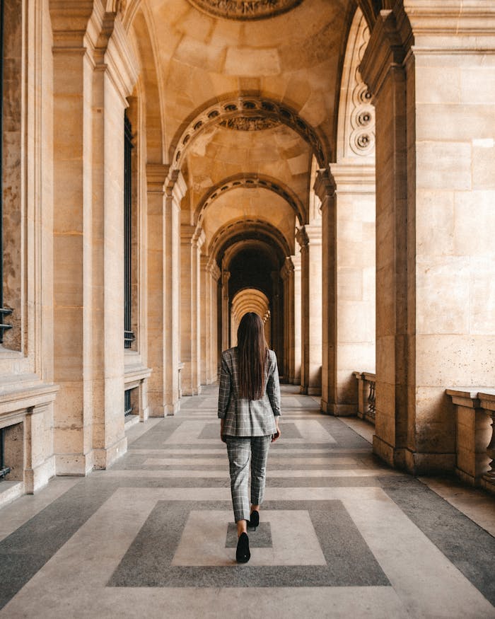 A woman in business attire walks through a historic hallway with arches and columns.